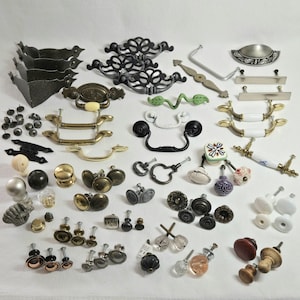 May include: A collection of various cabinet hardware, including knobs and pulls, in diverse styles and finishes. The assortment features metal, ceramic, and glass pieces in colors like black, gold, white, and clear. The hardware is designed for furniture and cabinetry.