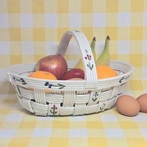 May include: A white ceramic basket with a woven texture and a single arched handle. The basket features hand-painted floral motifs in blue, green, and red along the rim and handle. It is filled with an apple, two bananas, and oranges.