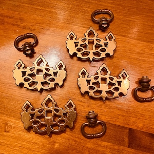 May include: A collection of antique brass drawer pulls and handles. The set includes five ornate, openwork escutcheons and four ring pulls. The hardware is arranged on a wooden surface, showcasing their aged, vintage aesthetic.