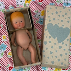 May include: A small, light-skinned baby doll with molded orange hair and painted facial features, positioned inside an open cardboard box. The box lid is decorated with a pattern of small blue hearts and two large, overlapping blue hearts.