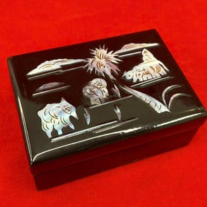 May include: A black rectangular box with a glossy finish. The box is decorated with inlaid mother-of-pearl designs depicting a landscape with animals, clouds, and a sun. The box sits on a red surface.