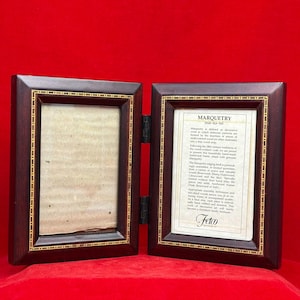 May include: A hinged, wooden photo frame with a dark brown finish and gold trim. One side is open, the other contains a printed description of "Marquetry". The frame is displayed against a red background.