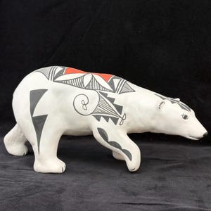May include: A ceramic bear figurine, painted white with black and red geometric designs. The bear is depicted in a walking pose, with detailed features and a glossy finish. The artwork is likely handcrafted.