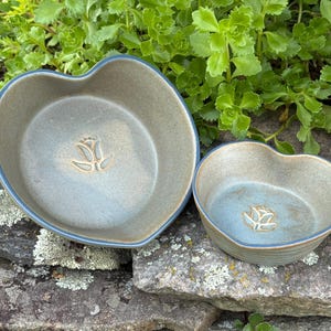 May include: Two heart-shaped ceramic bowls in a muted grey-blue glaze. Each bowl features a smooth, curved rim and a stamped floral motif at the center of the basin. The bowls are finished with a subtle blue edge along the top rim of the vessels.
