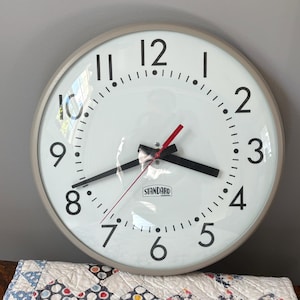 May include: A round wall clock with a light gray frame and a white face featuring black Arabic numerals from 1 to 12. The clock has black hour and minute hands, a thin red second hand, and the word STANDARD printed in a black box on the face.