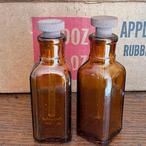 May include: Two amber-colored glass bottles with cork stoppers. The bottles have a rectangular shape and a long, thin tube inside. The background includes a red label with white text and a cardboard box.