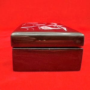 Op de afbeelding: Een rechthoekige, glanzend zwarte doos met een scharnierend deksel. Het deksel heeft een wit, ge&euml;tst ontwerp. De doos staat tegen een levendige rode achtergrond, wat een opvallend contrast cre&euml;ert. De doos lijkt van een hard materiaal te zijn gemaakt.