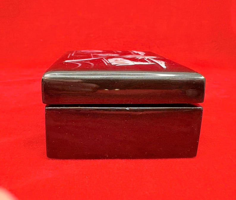 Op de afbeelding: Een rechthoekige, glanzend zwarte doos met een scharnierend deksel. Het deksel heeft een wit, ge&euml;tst ontwerp. De doos staat tegen een levendige rode achtergrond, wat een opvallend contrast cre&euml;ert. De doos lijkt van een hard materiaal te zijn gemaakt.