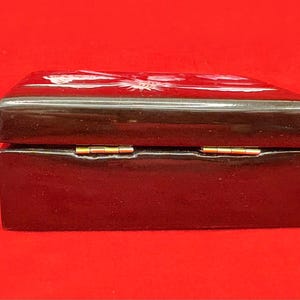 Op de afbeelding: Een rechthoekige, donkerbruine houten doos met een glanzende afwerking. De doos heeft een scharnierend deksel met goudkleurige metalen scharnieren. De bovenkant van de doos heeft een wit bloemenontwerp. De doos staat op een rode achtergrond.