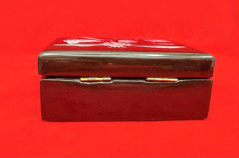 Op de afbeelding: Een rechthoekige, donkerbruine houten doos met een glanzende afwerking. De doos heeft een scharnierend deksel met goudkleurige metalen scharnieren. De bovenkant van de doos heeft een wit bloemenontwerp. De doos staat op een rode achtergrond.