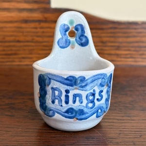 May include: A small, off-white ceramic wall-mounted holder with a rounded basin and a back tab featuring a circular hole for hanging. The piece is hand-painted with blue scrollwork patterns and the word "Rings" centered on the front surface.