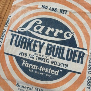 May include: Vintage feed sack with the text "Larro TURKEY BUILDER" in navy blue and white. The sack has orange and white concentric circles and the text "100 LBS. NET". The sack also has the text "Farm-tested" and "General Mills, Inc."