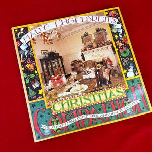 May include: A square hardcover book titled Mary Engelbreit's Christmas Companion. The cover features a central photograph of a decorated dining room with a fireplace, surrounded by a black border with colorful illustrations of holiday icons.