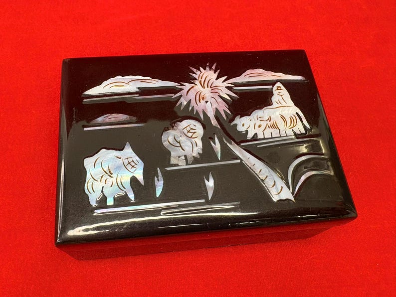 Op de afbeelding: Zwarte rechthoekige doos met een glanzende afwerking, versierd met een ingelegd parelmoer ontwerp. Het ontwerp toont een tropische sc&egrave;ne met palmbomen, wolken en gestileerde dieren. De doos staat tegen een rode achtergrond.