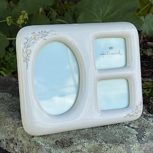 May include: A cream-colored ceramic picture frame with a crackle glaze finish. The frame features one large oval opening and two smaller square openings. Delicate floral accents are painted on the corners, and the Hallmark logo is printed.