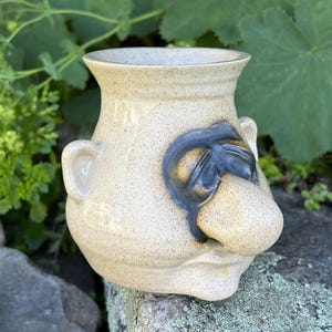 May include: A beige speckled ceramic mug featuring a sculpted face with a prominent nose, dark gray recessed eyes, and a small ear on the side. The mug has a wide, flared rim and a rounded base, crafted with a smooth, glossy finish throughout.