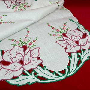 May include: White embroidered table runner with a scalloped edge. Features large pink and red floral designs with green and pink accents. The runner is on a red surface.