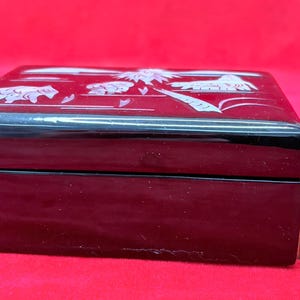 Op de afbeelding: Zwarte rechthoekige sieradendoos met een decoratief wit ontwerp op het deksel. De doos is ongeveer 5 cm breed. De doos staat op een rode ondergrond. De doos is gesloten.