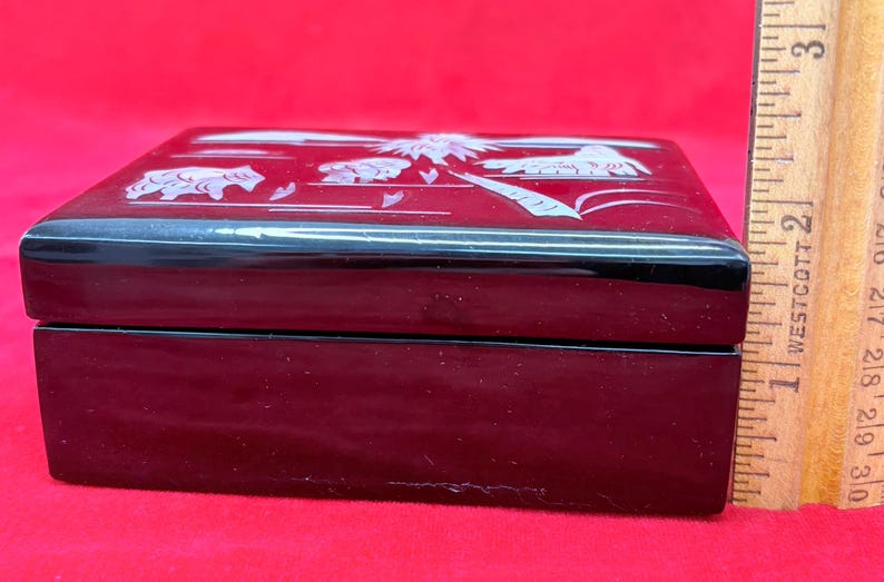 Op de afbeelding: Zwarte rechthoekige sieradendoos met een decoratief wit ontwerp op het deksel. De doos is ongeveer 5 cm breed. De doos staat op een rode ondergrond. De doos is gesloten.