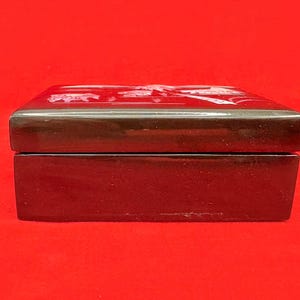 Op de afbeelding: Een rechthoekige, donkerbruine houten doos met een glanzende afwerking. De deksel heeft een wit bloemenontwerp. De doos staat tegen een felrode achtergrond, wat de rijke kleur en ingewikkelde details benadrukt. De doos wordt waarschijnlijk gebruikt voor sieraden of kleine aandenkens.