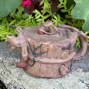May include: A brown ceramic teapot shaped like a tree stump with a textured bark surface. The lid features a small sculpted lizard figure. Additional sculpted details include a branch-like spout, a handle, and small lizard accents on the body.