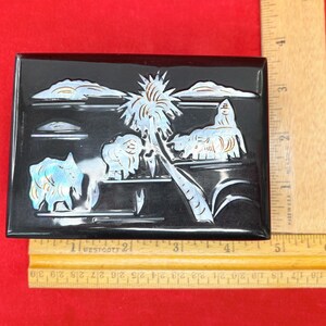 Op de afbeelding: Zwarte rechthoekige doos met een tropische sc&egrave;ne die in de bovenkant is gesneden. De sc&egrave;ne toont een palmboom, wolken en dieren, allemaal met iriserende accenten. Een liniaal staat naast de doos.