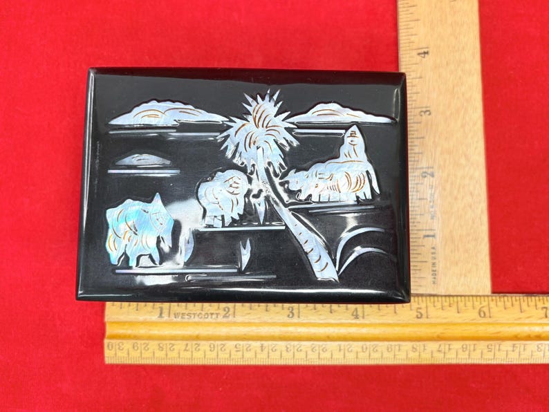 Op de afbeelding: Zwarte rechthoekige doos met een tropische sc&egrave;ne die in de bovenkant is gesneden. De sc&egrave;ne toont een palmboom, wolken en dieren, allemaal met iriserende accenten. Een liniaal staat naast de doos.