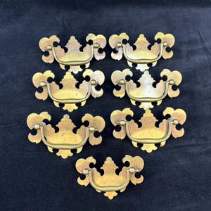 May include: Set of seven antique brass drawer pulls. Each pull features a decorative, ornate design with a curved handle and a detailed backplate. The brass has a warm, aged patina, suggesting vintage craftsmanship. Ideal for furniture restoration projects.