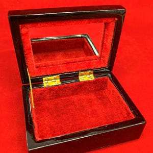 Op de afbeelding: Open zwarte sieradendoos met een rode fluwelen binnenkant en een spiegeldeksel. De doos is rechthoekig met goudkleurige scharnieren. De binnenkant is felrood, in contrast met de glanzende zwarte buitenkant.