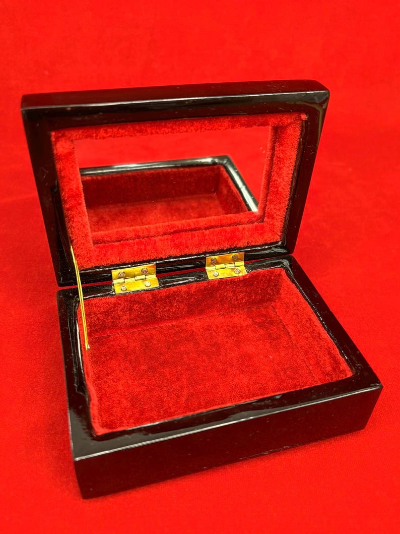 Op de afbeelding: Open zwarte sieradendoos met een rode fluwelen binnenkant en een spiegeldeksel. De doos is rechthoekig met goudkleurige scharnieren. De binnenkant is felrood, in contrast met de glanzende zwarte buitenkant.