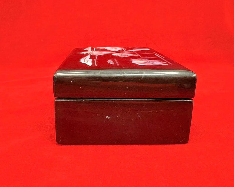 Op de afbeelding: Een rechthoekige, donkerbruine doos met een glanzende afwerking. De bovenkant van de doos heeft een wit bloemenontwerp. De doos is gesloten en staat tegen een rode achtergrond. De doos lijkt van hout te zijn.