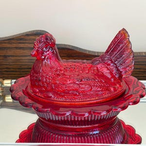 May include: A ruby red glass hen-shaped covered dish. The dish features a detailed hen design with textured feathers and a scalloped base. The dish is displayed on a mirrored tray, showcasing its vibrant color and intricate details. A decorative kitchenware item.