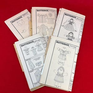 May include: A collection of vintage Butterick sewing patterns, featuring illustrations of dolls and clothing designs. The patterns are printed on white paper with black ink, showcasing various garment styles. The patterns are arranged on a red surface.