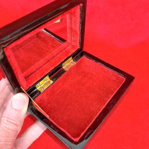 Op de afbeelding: Open zwarte gelakte sieradendoos met een rode fluwelen binnenkant en een kleine spiegel aan de binnenkant van het deksel. De doos is rechthoekig met goudkleurige scharnieren.