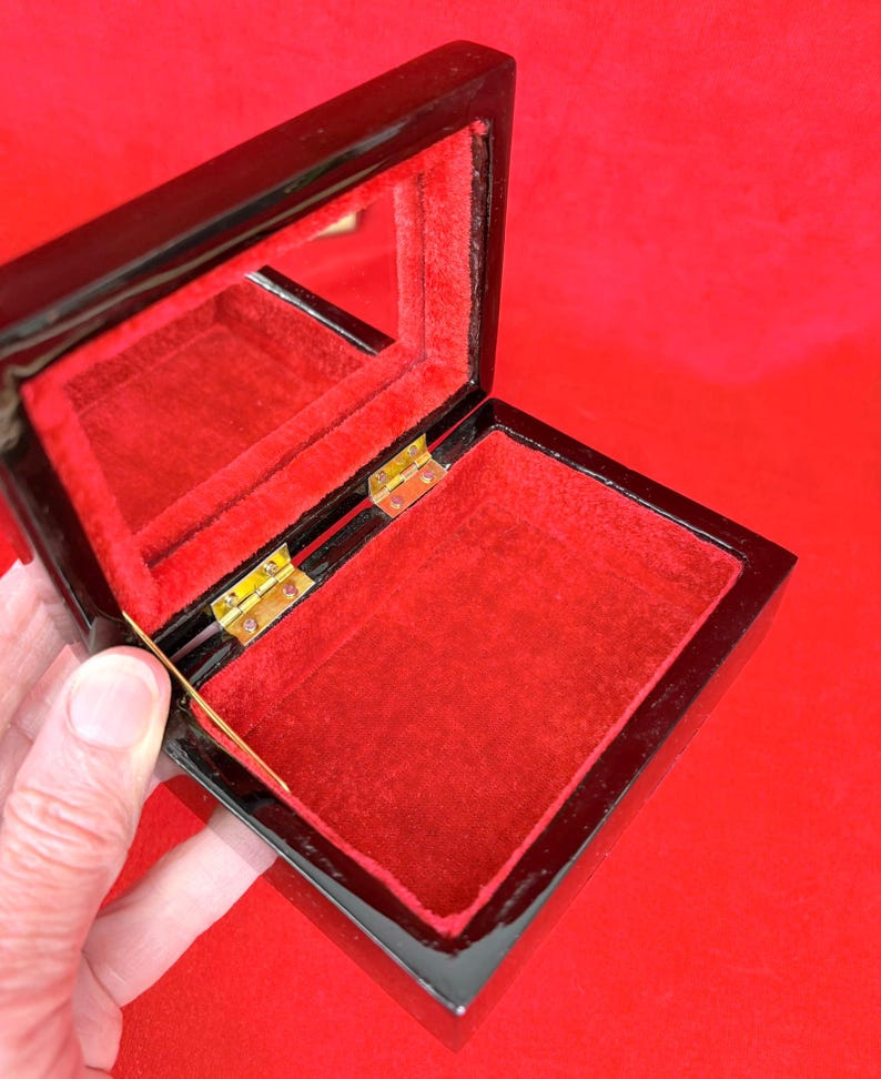 Op de afbeelding: Open zwarte gelakte sieradendoos met een rode fluwelen binnenkant en een kleine spiegel aan de binnenkant van het deksel. De doos is rechthoekig met goudkleurige scharnieren.