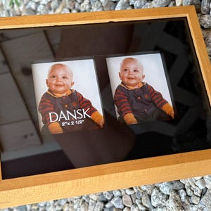 May include: A rectangular light wood picture frame with a black mat featuring two openings for photos. The left opening contains a photo of a baby and the text DANSK 2" x 2 1/2". The right opening contains a photo of the same baby.