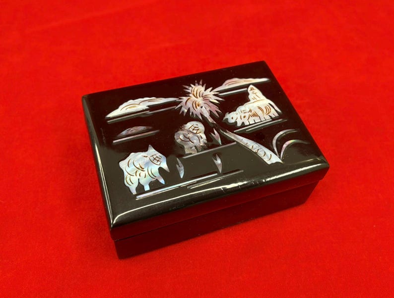 Op de afbeelding: Een zwarte rechthoekige doos met een glanzende afwerking. De doos is versierd met ingelegde parelmoer ontwerpen die een landschap met dieren, wolken en een zon uitbeelden. De doos staat op een rode ondergrond.