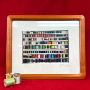 May include: A framed artwork featuring a collection of colorful thread spools arranged on shelves. The wooden frame surrounds a white mat, and the background is a vibrant red. Several spools of thread are in front of the frame.