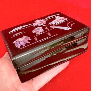 Op de afbeelding: Een rechthoekige, donkerbruine sieradendoos met een glanzende afwerking. De deksel is versierd met ingelegde parelmoer ontwerpen van dieren en bloemmotieven. De doos wordt in een hand gehouden tegen een rode achtergrond.