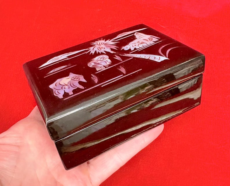 Op de afbeelding: Een rechthoekige, donkerbruine sieradendoos met een glanzende afwerking. De deksel is versierd met ingelegde parelmoer ontwerpen van dieren en bloemmotieven. De doos wordt in een hand gehouden tegen een rode achtergrond.