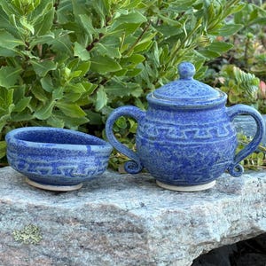 May include: A blue ceramic teapot with a matching sugar bowl. The teapot has a lid and a handle. Both pieces have a textured surface and a white base. The set is placed on a stone surface with green foliage in the background.
