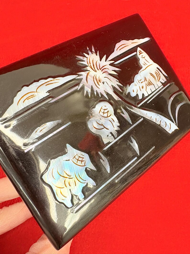 Op de afbeelding: Een rechthoekige zwarte doos met ingelegde parelmoer ontwerpen. De doos heeft een landschap met een zonnestraal, wolken en gestileerde vormen. De iriserende details cre&euml;ren een opvallend contrast met de donkere achtergrond.