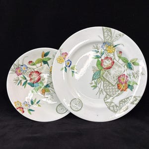 May include: Two white ceramic plates with floral designs. The plates feature colorful flowers, including red, yellow, and blue blooms, with green leaves and stems. The plates have a decorative border and are set against a black background.