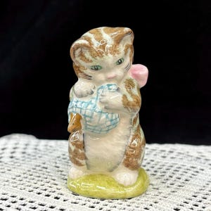 May include: A ceramic figurine of a kitten holding a blue and white basket with a small brown mouse. The kitten has brown and white striped fur, green eyes, and a pink bow. The figurine is on a green base.