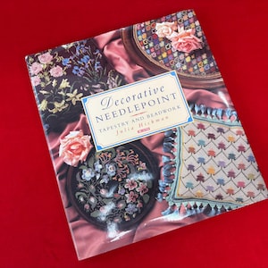 May include: A book titled "Decorative Needlepoint Tapestry and Beadwork" by Julia Hickman. The cover features images of needlepoint and beadwork designs, including floral patterns and a framed piece with a blue tassel border. The background is a red surface.
