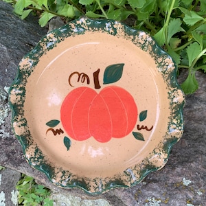 May include: A round ceramic pie dish with a tan speckled base and a ruffled, dark green sponge-painted rim. The center features a hand-painted orange pumpkin illustration with brown curly vines and small green leaves on a smooth, glazed surface.