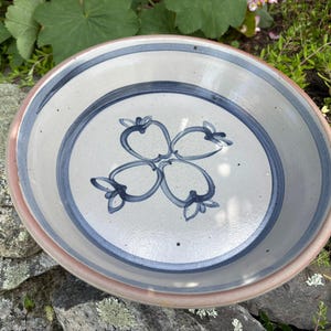 May include: A round ceramic bowl with a light beige base and a smooth, glossy finish. The interior features a hand-painted blue design of four stylized fruit shapes arranged in a central cluster, surrounded by two concentric blue circular bands.