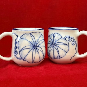 May include: Two white ceramic mugs with blue floral designs. Each mug features a large, stylized blue flower on the front and a blue rim. The mugs have white handles and are set against a red background.