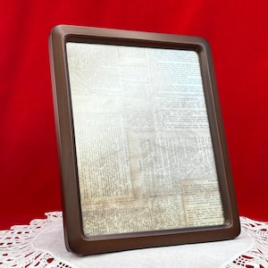 May include: A rectangular picture frame with a smooth, dark brown border. The frame contains a decorative insert featuring a collage of vintage-style newspaper clippings with black text on a light, aged, off-white paper background.