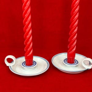 May include: Two red, twisted taper candles sit in white ceramic candle holders with blue and white detailing. Each holder has a small handle. The background is a solid red.